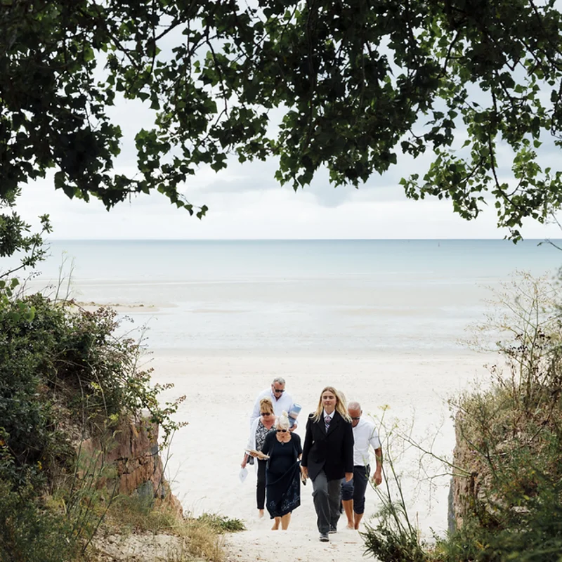 Funeral director and family walking together along a sandy beach path.