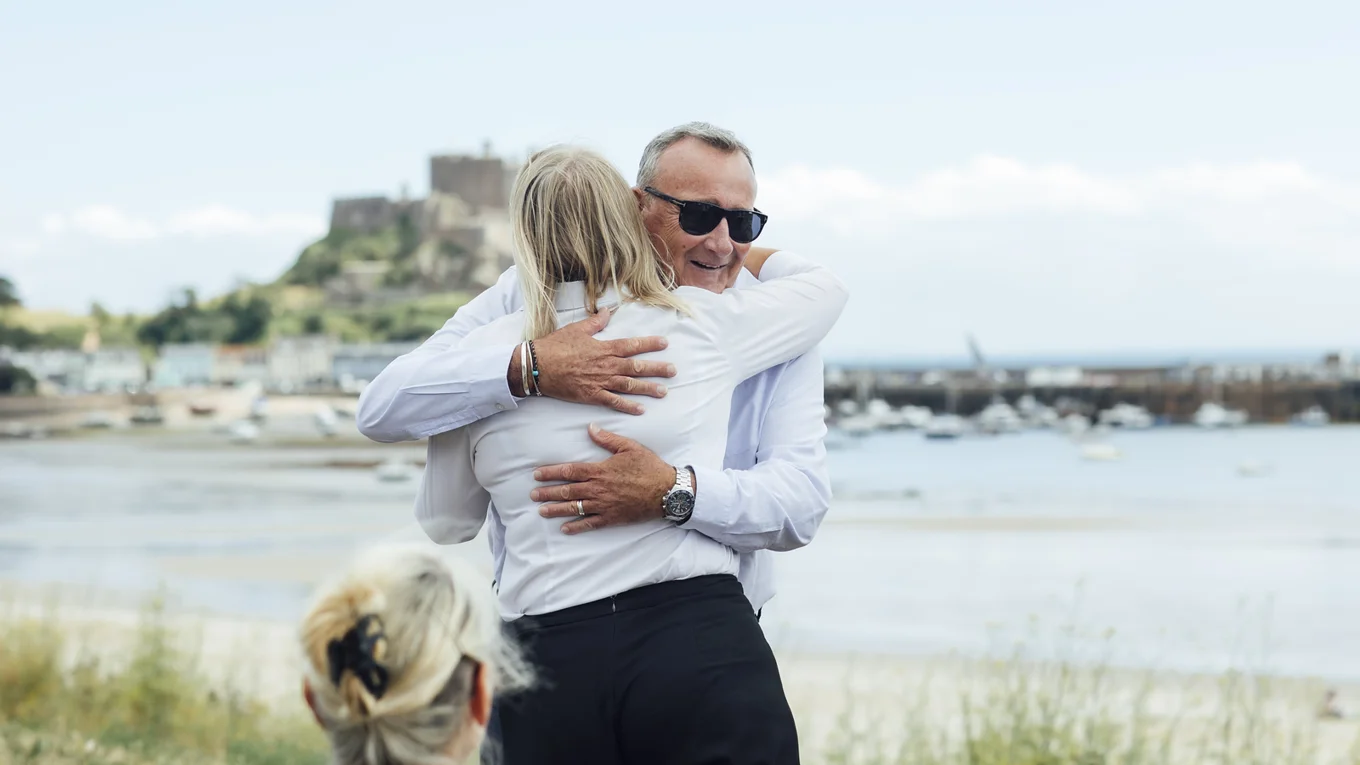 A man embraces a woman by the harbour, with Gorey Castle in the background.